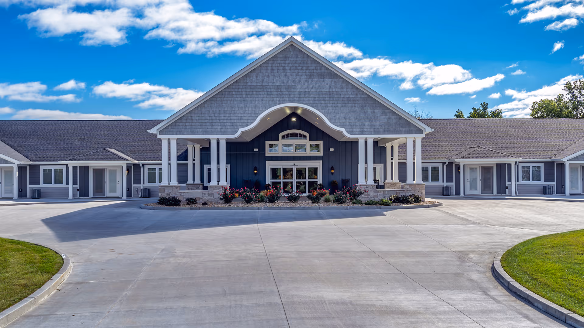 Front exterior view of Villas of Holly Brook Assisted Living & Memory Care building in Springfield, IL, featuring a large covered entrance with white pillars, gray siding, and a landscaped area with flowers in front. The sky is blue with scattered clouds.