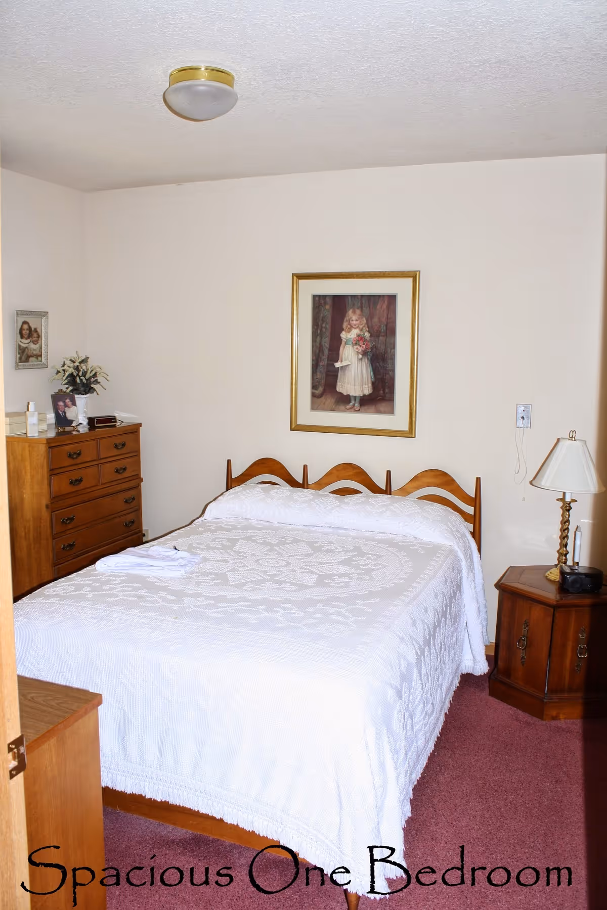 Neat single bedroom with a bed covered by a white bedspread, wooden dressers, framed wall art, and a bedside lamp.
