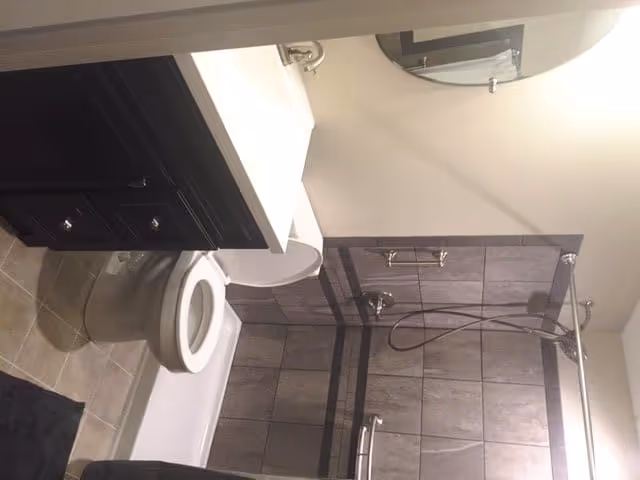 Bathroom with a white toilet, a dark wood vanity with a white countertop and sink, a large round mirror above the sink, and a bathtub with a tiled shower surround featuring gray tiles and a mounted showerhead with a handheld attachment.