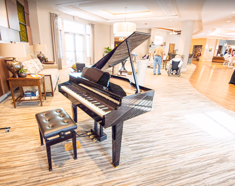 A glossy black grand piano and bench sit in a bright, carpeted senior living facility lounge with sofas, lamps, and residents in the background.