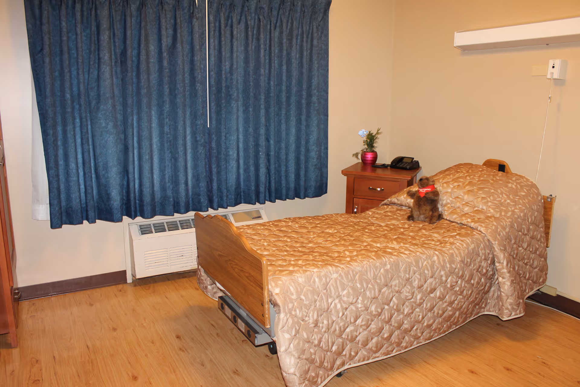 A small bedroom in a healthcare facility with a single bed covered in a beige quilted bedspread. There is a small stuffed animal with a red bow on the bed. Next to the bed is a wooden nightstand with a pink vase holding a small plant and a black telephone. The room has wooden flooring, beige walls, a blue curtain covering the window, and an air conditioning unit below the window.