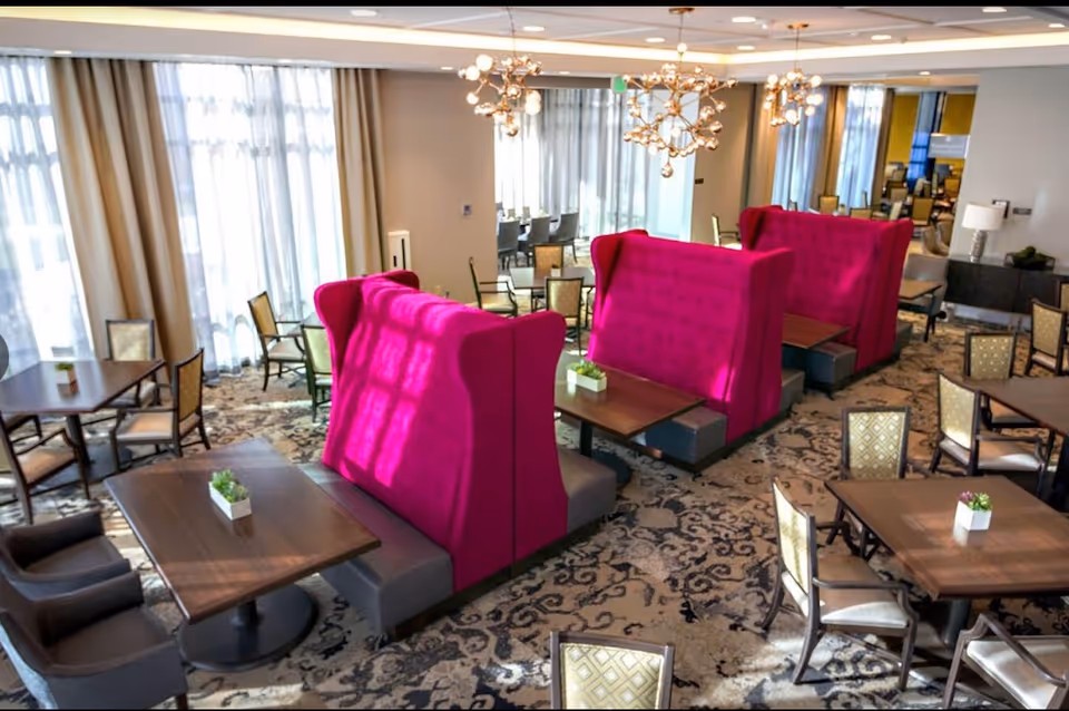 Bright communal dining/lounge area with magenta high-backed booth seating, wooden tables and chairs, and large windows.