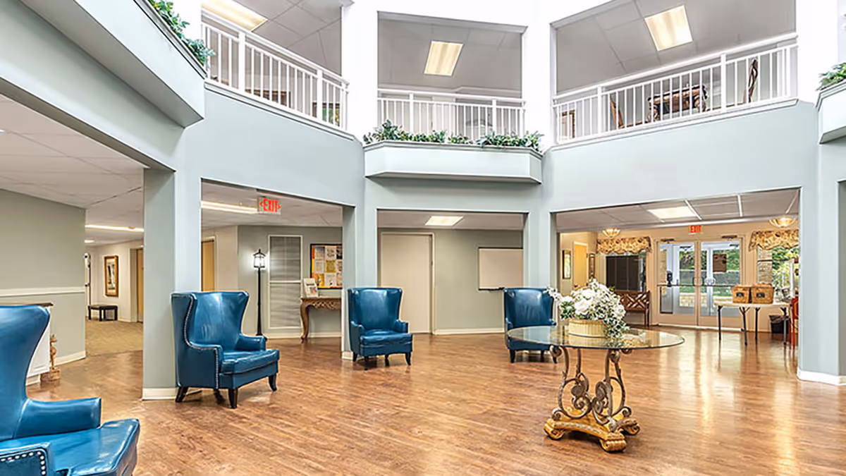 Spacious and well-lit common area in a senior living facility with high ceilings, blue armchairs arranged around the room, a round glass table with a floral centerpiece, and a second-floor balcony with white railings overlooking the area.