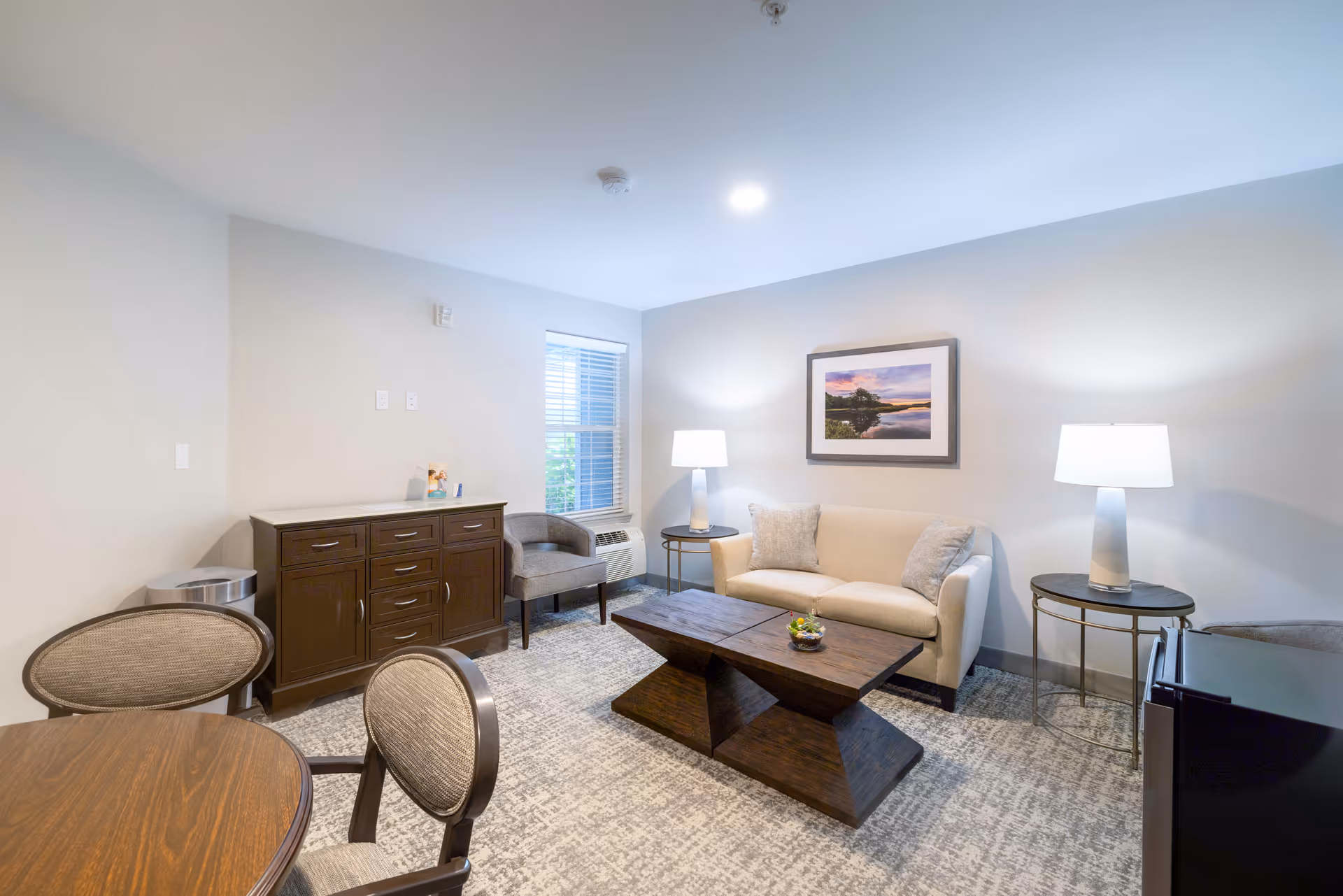 A cozy living room with a beige sofa, two side tables with lamps, a dark wooden coffee table, a dark wooden cabinet, two upholstered chairs, a round wooden dining table with two chairs, a window with blinds, and a framed landscape picture on the wall.