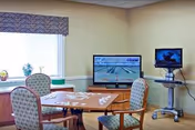 A senior living common room with a wooden activity table and four upholstered chairs, two TVs, and a window with a valance.