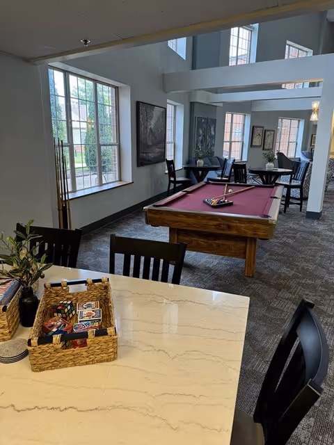 Interior view of a common area in a senior living facility featuring a pool table with a red felt top, several black chairs and tables, large windows letting in natural light, and a countertop with baskets containing playing cards and a Rubik's cube.