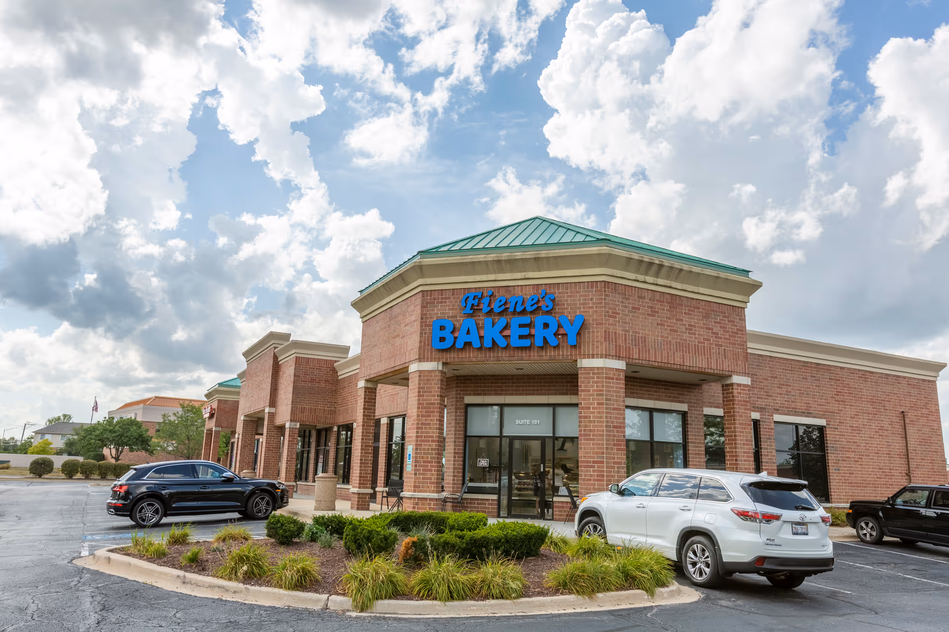 Brick storefront of Fiene's Bakery with a green roof and parked cars in front under a cloudy sky.