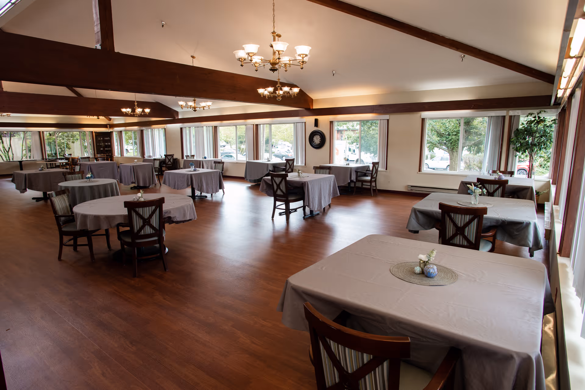 Spacious dining room with multiple tables covered in gray tablecloths, each set with small decorative centerpieces. Wooden chairs surround the tables. Large windows along the walls let in natural light and offer views of trees and parked cars outside. The room features wooden beams on the ceiling and elegant chandeliers hanging down.