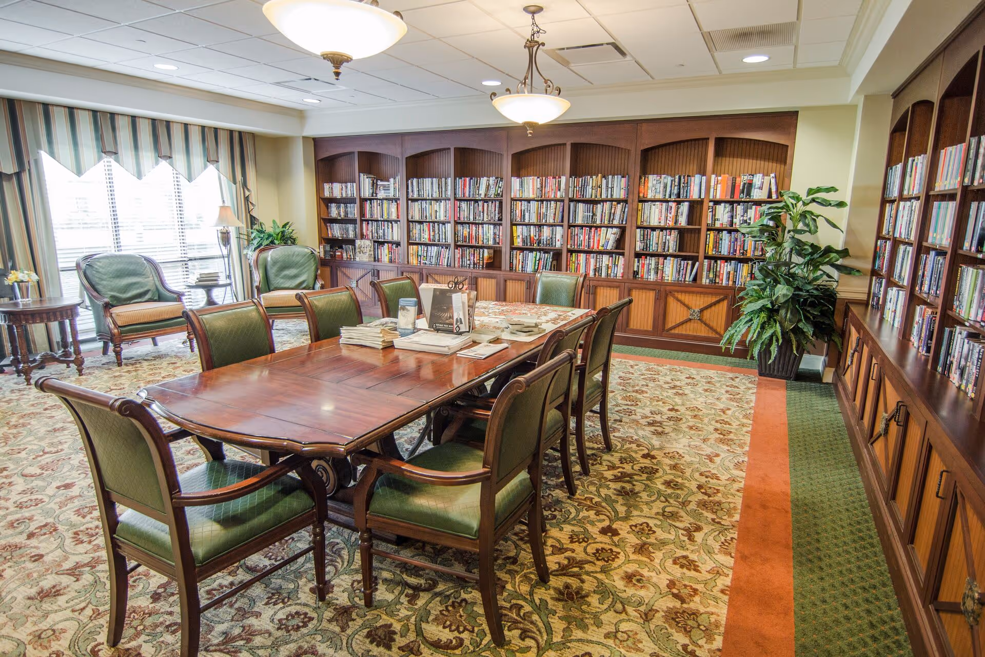 A well-lit library room with wooden bookshelves filled with books lining the walls. In the center, there is a large wooden table surrounded by green upholstered chairs. The room has patterned carpet flooring, two ceiling lights, and large windows with striped curtains allowing natural light to enter. There are also two green armchairs near the windows and a potted plant in the corner.