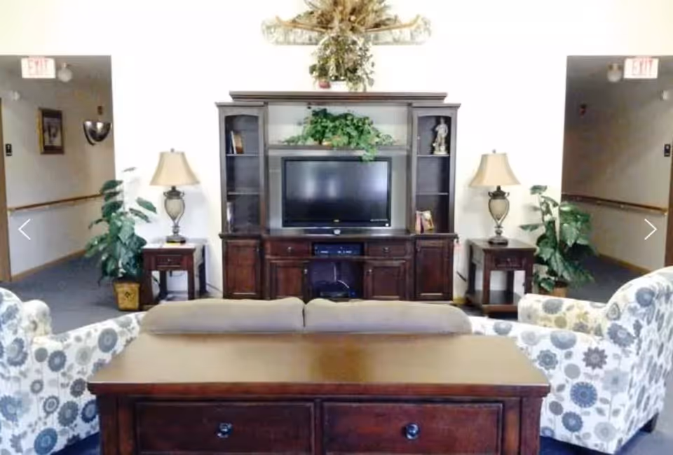 A cozy living room area with a wooden entertainment center holding a flat-screen TV, flanked by two table lamps on side tables. There are two patterned armchairs and a beige sofa facing the TV. Green plants and decorative items are placed around the room, with hallways visible on either side.