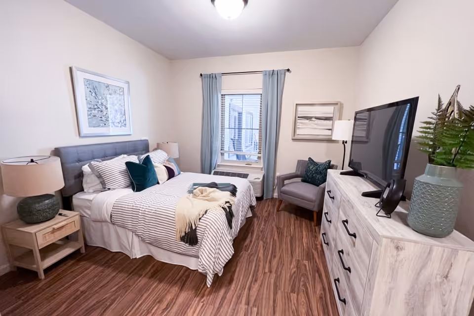 A cozy bedroom with a bed featuring striped bedding and multiple pillows, a wooden nightstand with a lamp, a window with light blue curtains, a gray armchair with a decorative pillow, a white dresser with a flat-screen TV on top, and a green vase with plants. The room has wood flooring and light-colored walls with framed artwork.
