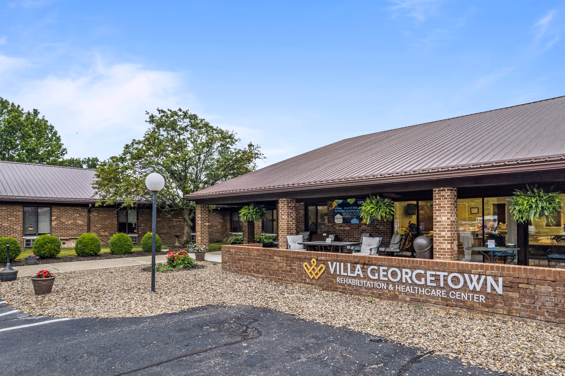 Exterior view of Villa Georgetown Rehabilitation and Healthcare Center showing a brick building with a covered patio area furnished with chairs and tables. There are hanging plants and a sign on a low brick wall displaying the facility's name. The surrounding area includes a parking lot, landscaping with bushes, a tree, and flower pots under a clear blue sky.