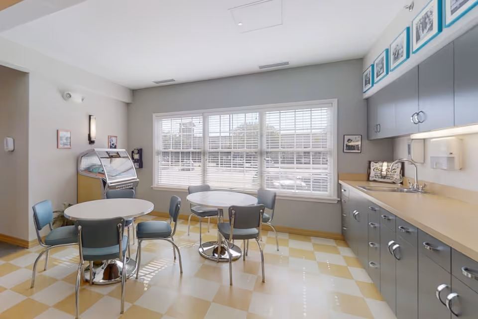 A bright room with large windows covered by white blinds, featuring two round tables each surrounded by four blue chairs. The room has a yellow and white checkered floor, gray cabinets with a countertop and sink on the right side, and a vintage jukebox against the left wall. Several framed pictures and a wall-mounted phone are also visible.