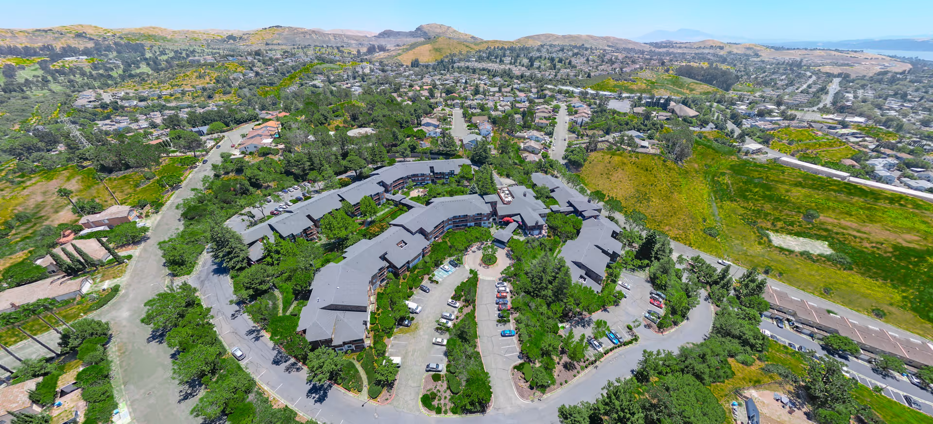 Aerial view of Cogir of Vallejo Hills Senior Living facility surrounded by greenery, parking lots, and residential neighborhoods with hills in the background under a clear blue sky.