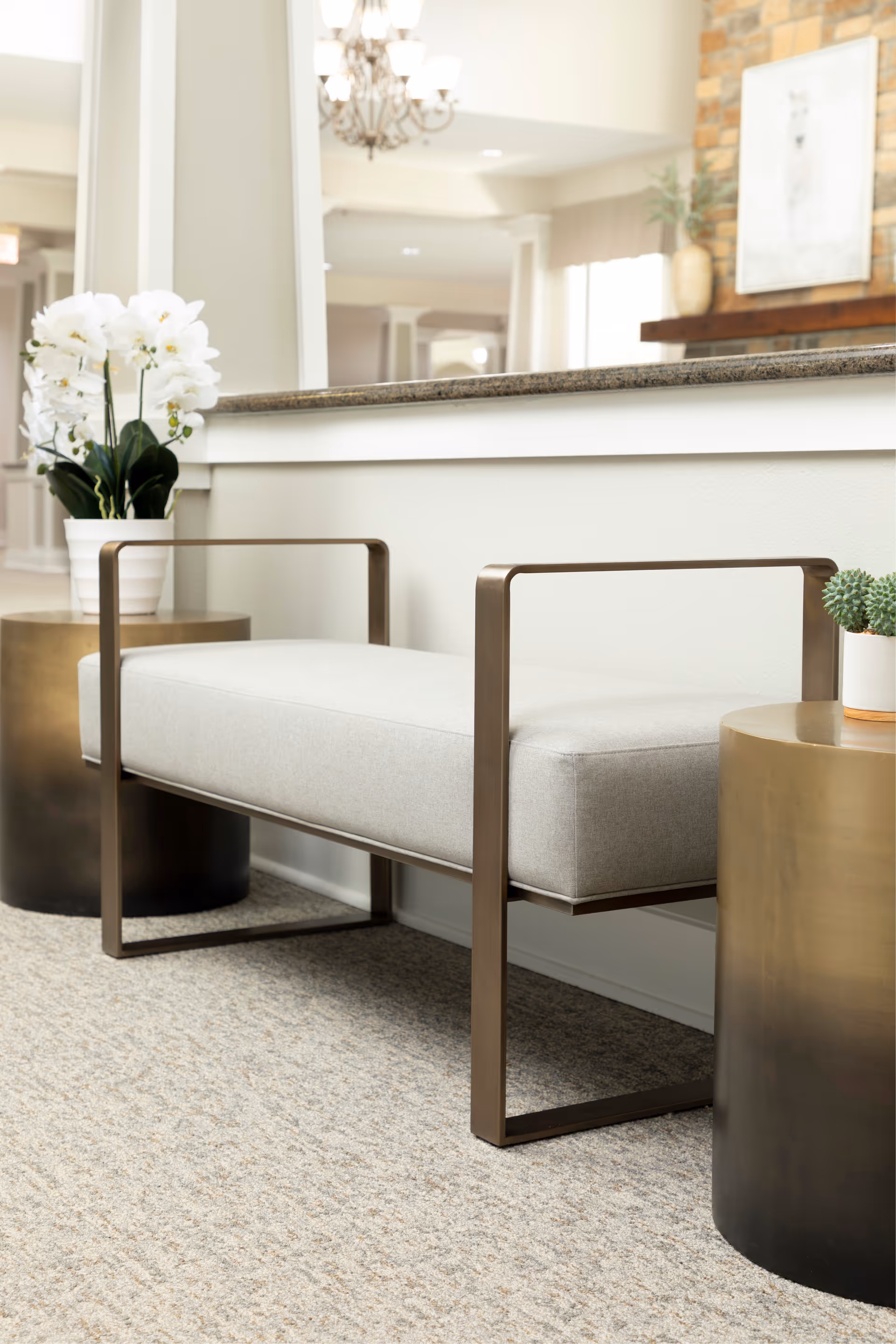 A modern seating area with a light gray cushioned bench with metal armrests, flanked by two round metallic side tables. One side table holds a white pot with white orchids, and the other has a small green plant. The background shows a partial view of a room with a chandelier and a stone fireplace with a framed artwork above it.