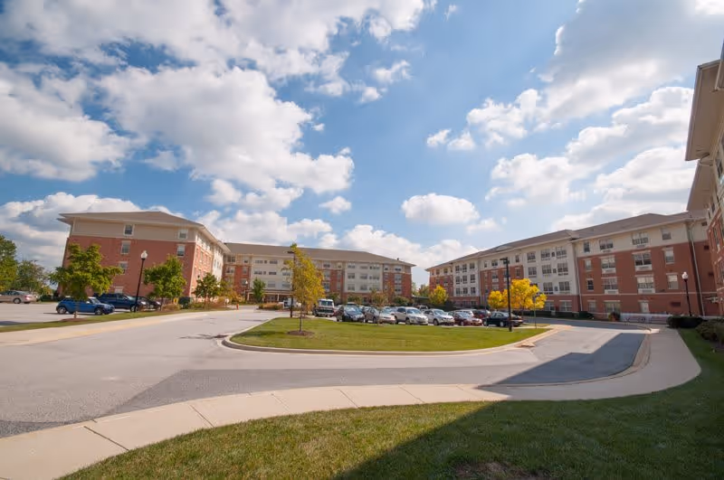 Exterior view of Ednor Apartments II showing multiple connected brick and beige buildings surrounding a parking lot with several cars parked. The sky is partly cloudy with blue patches visible.