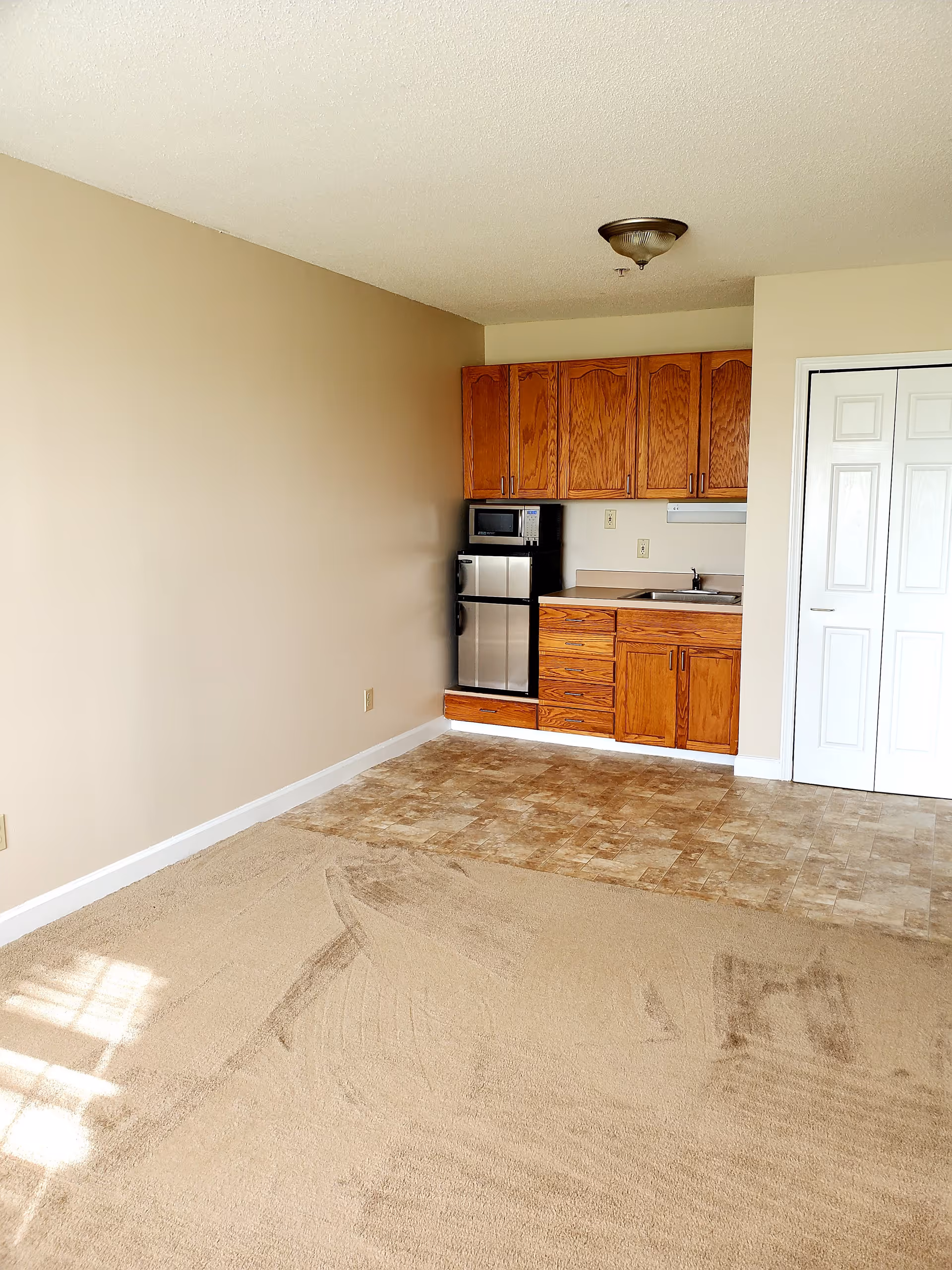 A small kitchenette area with wooden cabinets, a microwave, a mini refrigerator, and a sink. The floor transitions from carpet to tile near the kitchenette. There is a white double-door closet to the right and beige walls throughout the room.