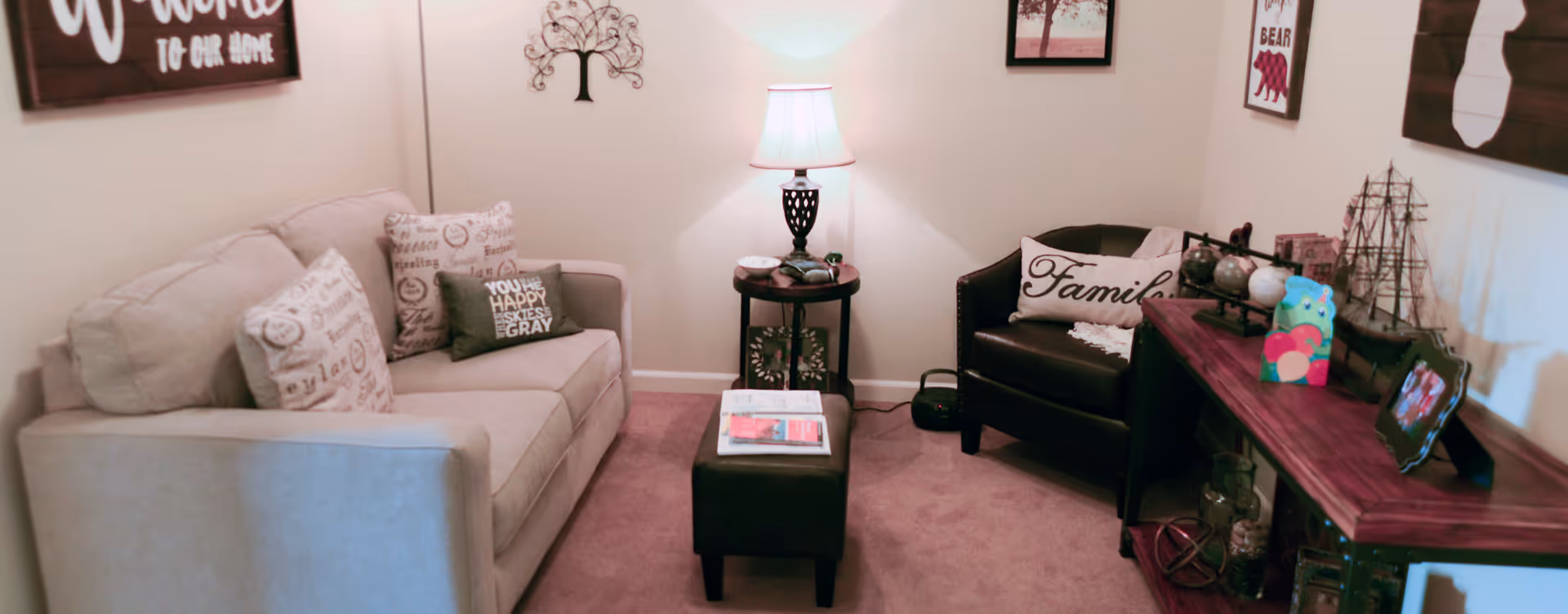 A cozy living room with a beige sofa adorned with decorative pillows, a black armchair with a 'Family' pillow, a small black ottoman, and a wooden console table displaying various decorative items including a model ship, globes, and framed photos. A table lamp on a round side table illuminates the space, and wall art decorates the walls.
