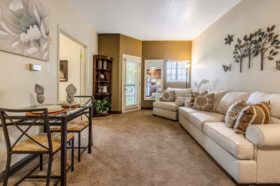 Carpeted living room with a beige sofa and armchair, decorative wall art, a small glass dining table, and a corner shelving unit.
