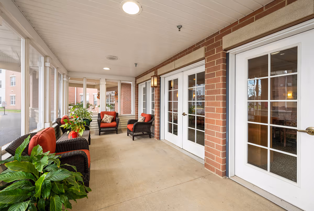 A covered outdoor seating area with wicker chairs featuring red cushions and decorative pillows. There are several green plants placed around the space. The area is enclosed with large windows and has brick walls with white-framed glass doors leading inside.