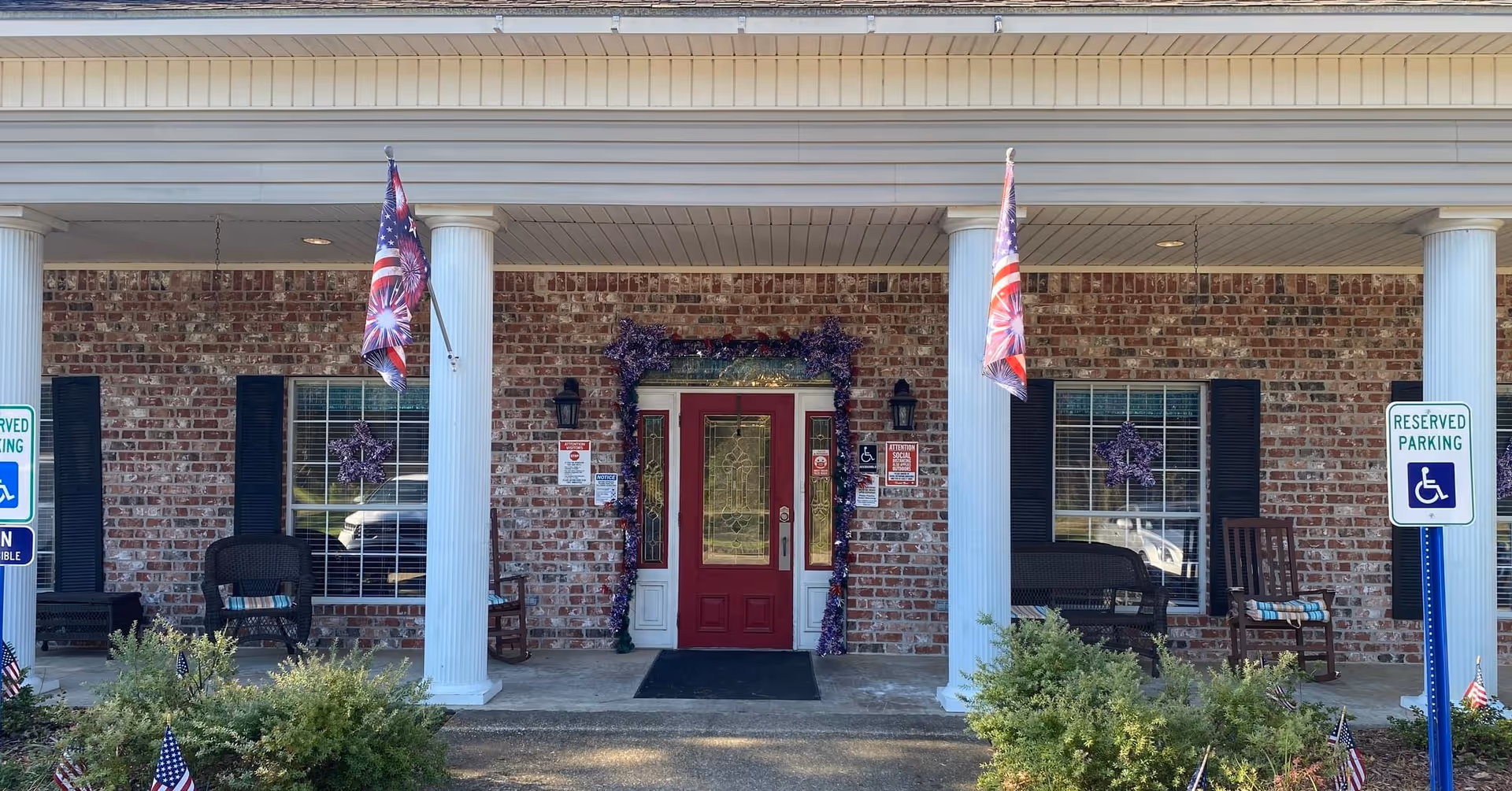 Front entrance of a brick building with white columns and a red door decorated with purple garland. Two American flags are hanging on either side of the door. There are two windows with black shutters and purple star decorations. Several chairs are placed on the porch, and there are reserved parking signs for handicapped parking in front of the building.