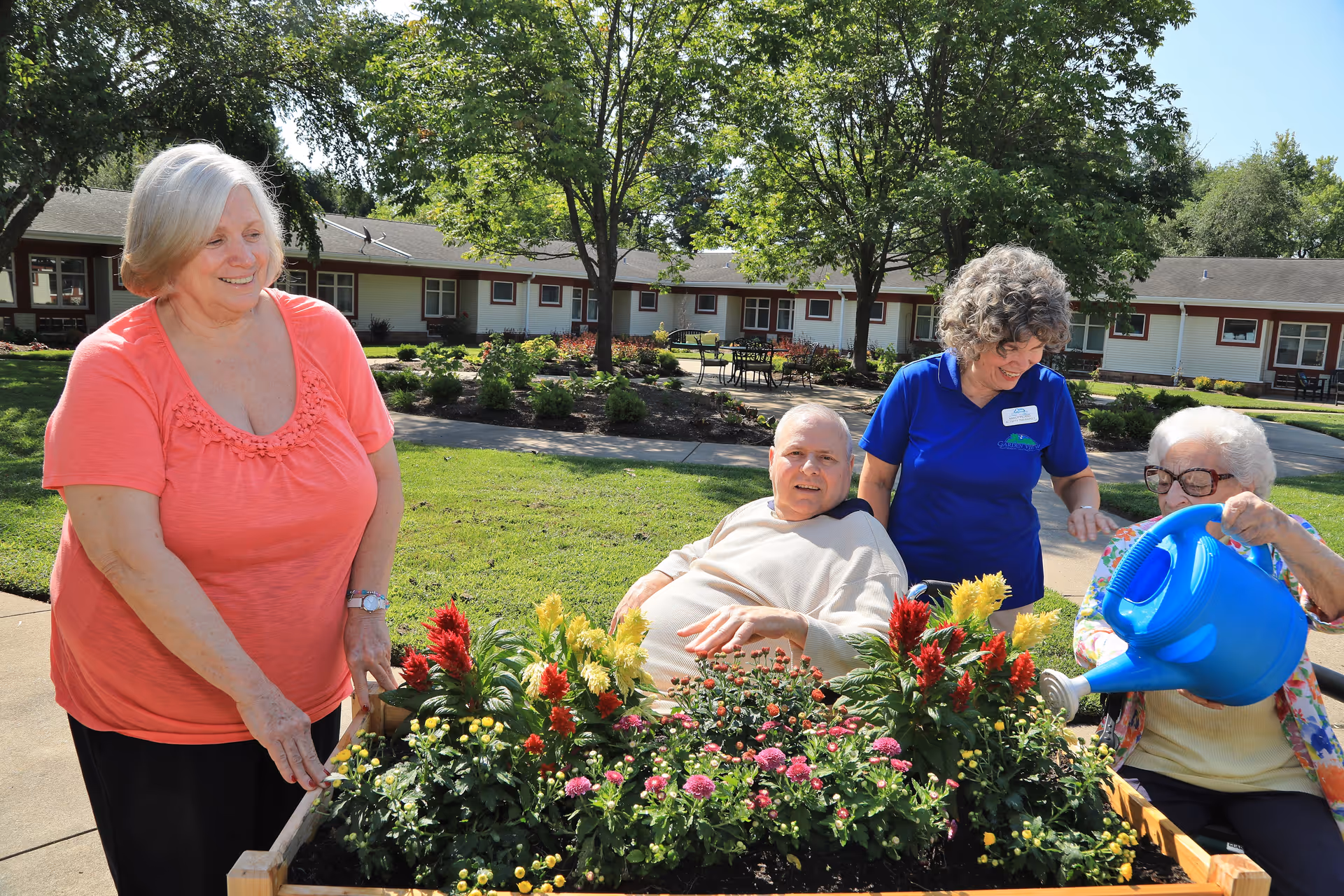 Four elderly individuals enjoying gardening outdoors at a senior living facility. One woman in a coral shirt is standing and smiling, a man in a wheelchair is seated near a raised garden bed filled with colorful flowers, a woman in a blue shirt is standing behind the man, and another woman is watering the plants with a blue watering can. The background shows a well-maintained garden area with trees and a single-story building.