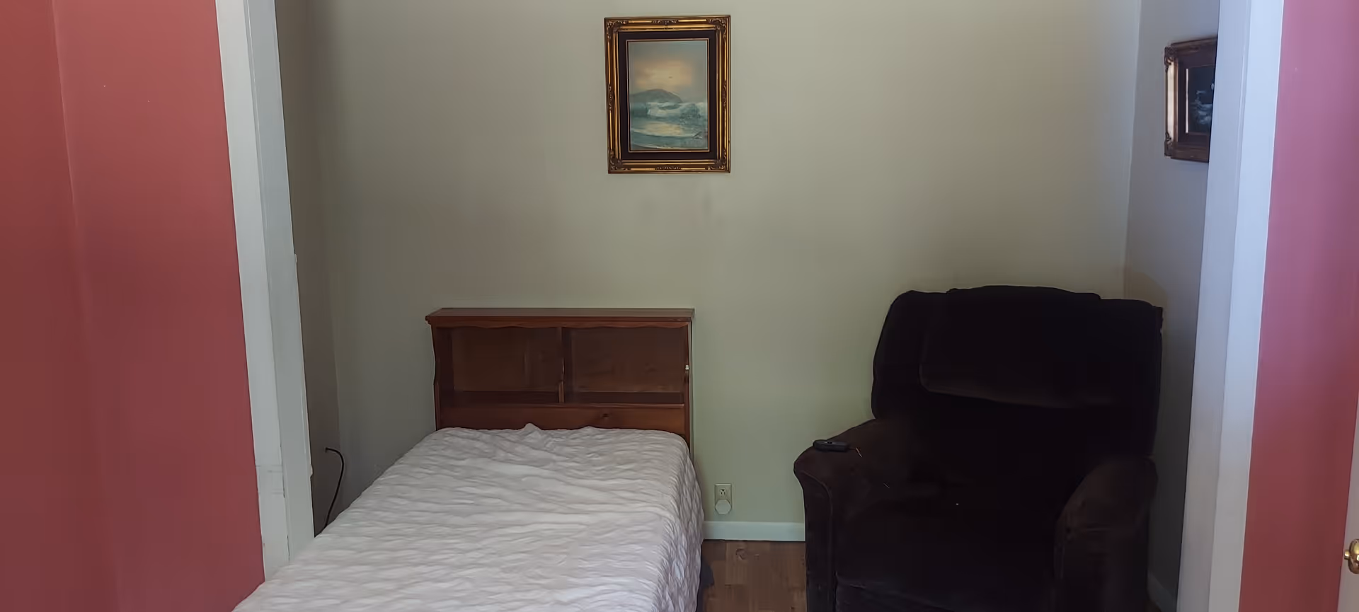 A small bedroom with a single bed covered with a white quilt on the left side and a dark upholstered armchair on the right side. There is a framed painting hanging on the beige wall above the bed. The walls on either side of the room are painted red.