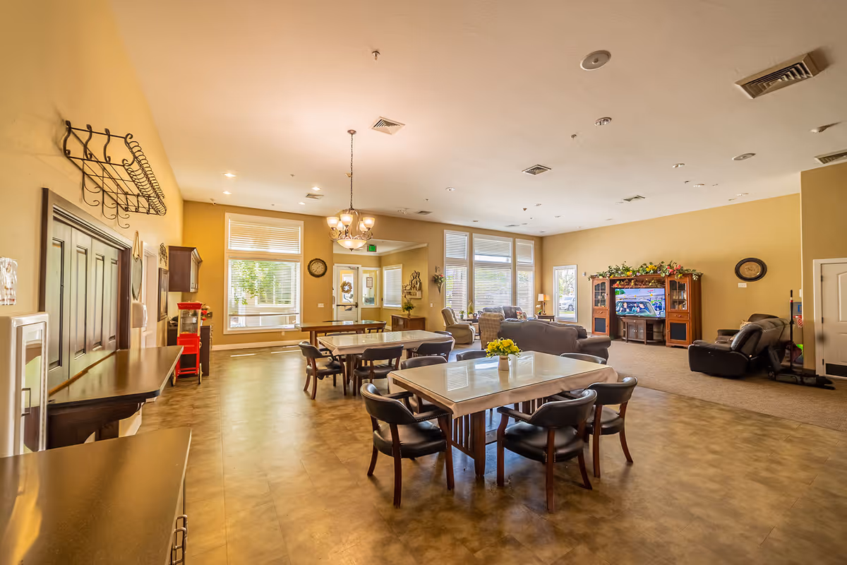 A spacious senior living common area with multiple tables and chairs in the foreground, a seating area with sofas and recliners facing a television in a wooden entertainment center, large windows letting in natural light, and warm beige walls. The room has a chandelier, wall clock, and some decorative plants.