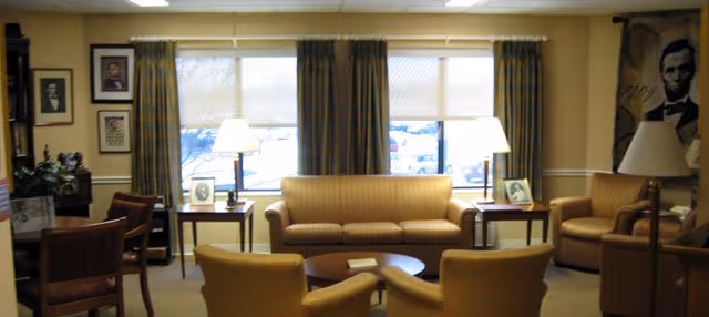 A cozy living room with a beige sofa, two armchairs, a round wooden coffee table, two side tables with lamps, framed pictures on the walls, and large windows with curtains letting in natural light.