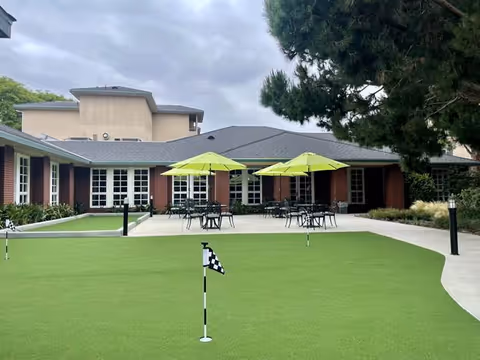 Outdoor patio area at Carmel Village Retirement Community featuring a putting green with small flags, several tables with green umbrellas, and a building with large windows and a brick exterior under a cloudy sky.