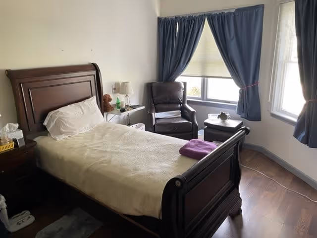A bedroom with a wooden bed frame and a beige blanket, a white pillow, a purple towel folded on the bed, a bedside table with a lamp and some items, a dark brown armchair near a window with blue curtains, and wooden flooring.