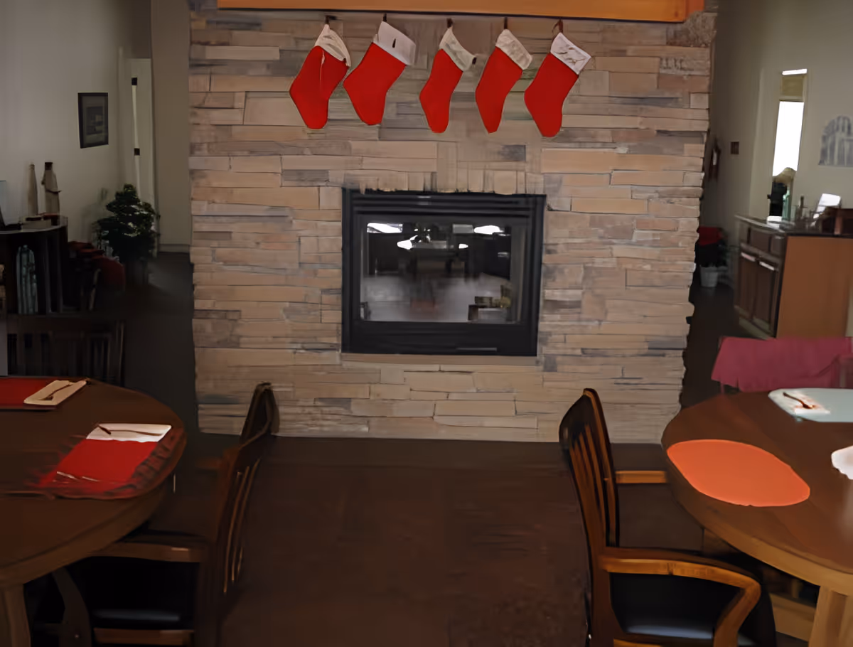 Interior view of a dining area with a stone fireplace in the center. Five red Christmas stockings are hanging above the fireplace. Two wooden dining tables with chairs are positioned on either side of the fireplace, each set with placemats and napkins. The room has a warm, cozy atmosphere with holiday decorations.