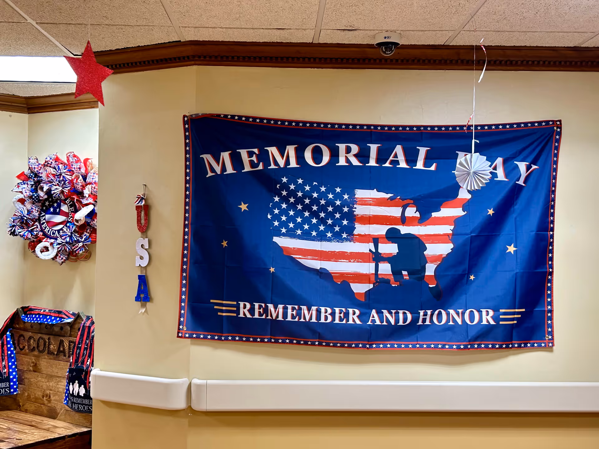Interior wall decorated with a large Memorial Day banner featuring a silhouette of a soldier kneeling in front of a cross overlaid on a U.S. flag map, with the words 'MEMORIAL DAY REMEMBER AND HONOR'. To the left, there is a patriotic wreath and hanging letters spelling 'USA'. A red star decoration hangs from the ceiling.