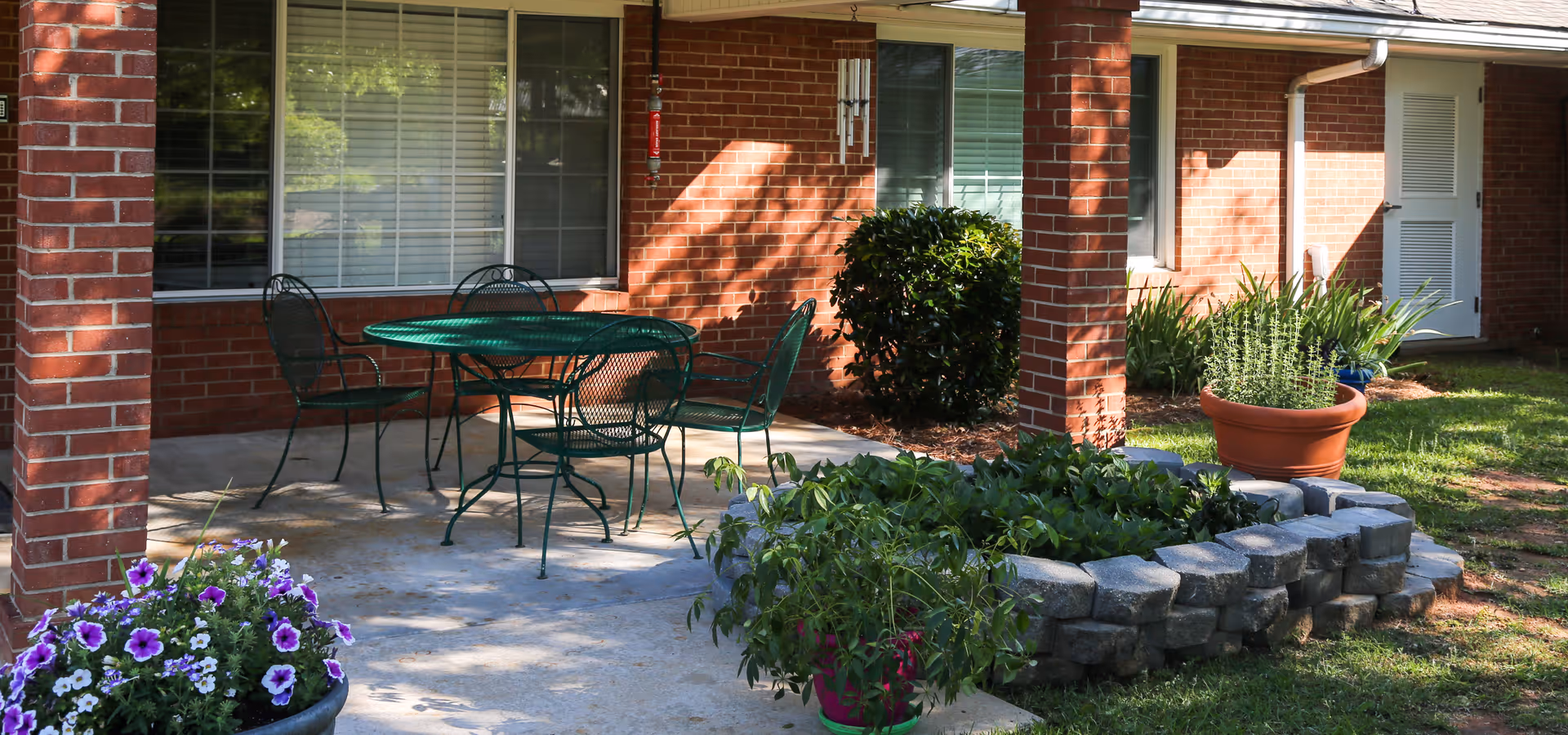 Covered brick patio with a green metal table and chairs, potted flowers, and a small raised stone planter in front of a red-brick building.