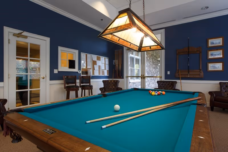 Interior room with a pool table in the center, two pool cues resting on the table, a rack with pool cues on the wall, several chairs around the room, a bulletin board on a blue wall, and a hanging light fixture above the pool table.
