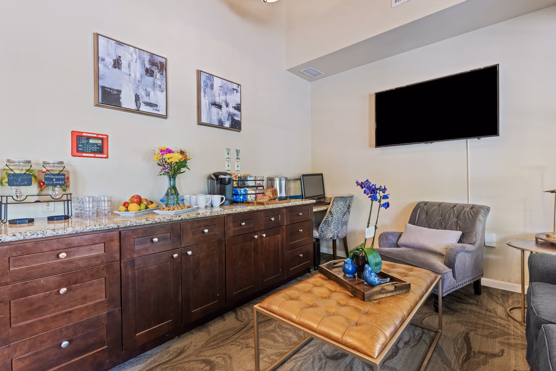 A cozy interior room with a long wooden cabinet topped with granite, holding jars of infused water, a plate of fruit, a vase with flowers, coffee cups, and a coffee maker. There is a small desk with a chair and a laptop in the corner. In the foreground, a tufted leather ottoman with a tray holding decorative items sits in front of a gray armchair with a pillow. A flat-screen TV is mounted on the wall above the armchair, and two abstract paintings hang on the wall above the cabinet.
