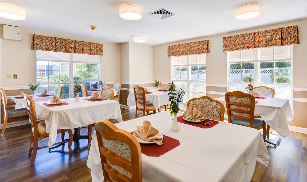 A bright dining room with several tables covered in white tablecloths and set with folded beige napkins on red placemats. Each table has wooden chairs with floral upholstery. Large windows with patterned valances let in natural light and show greenery outside. The room has wooden flooring and ceiling lights.