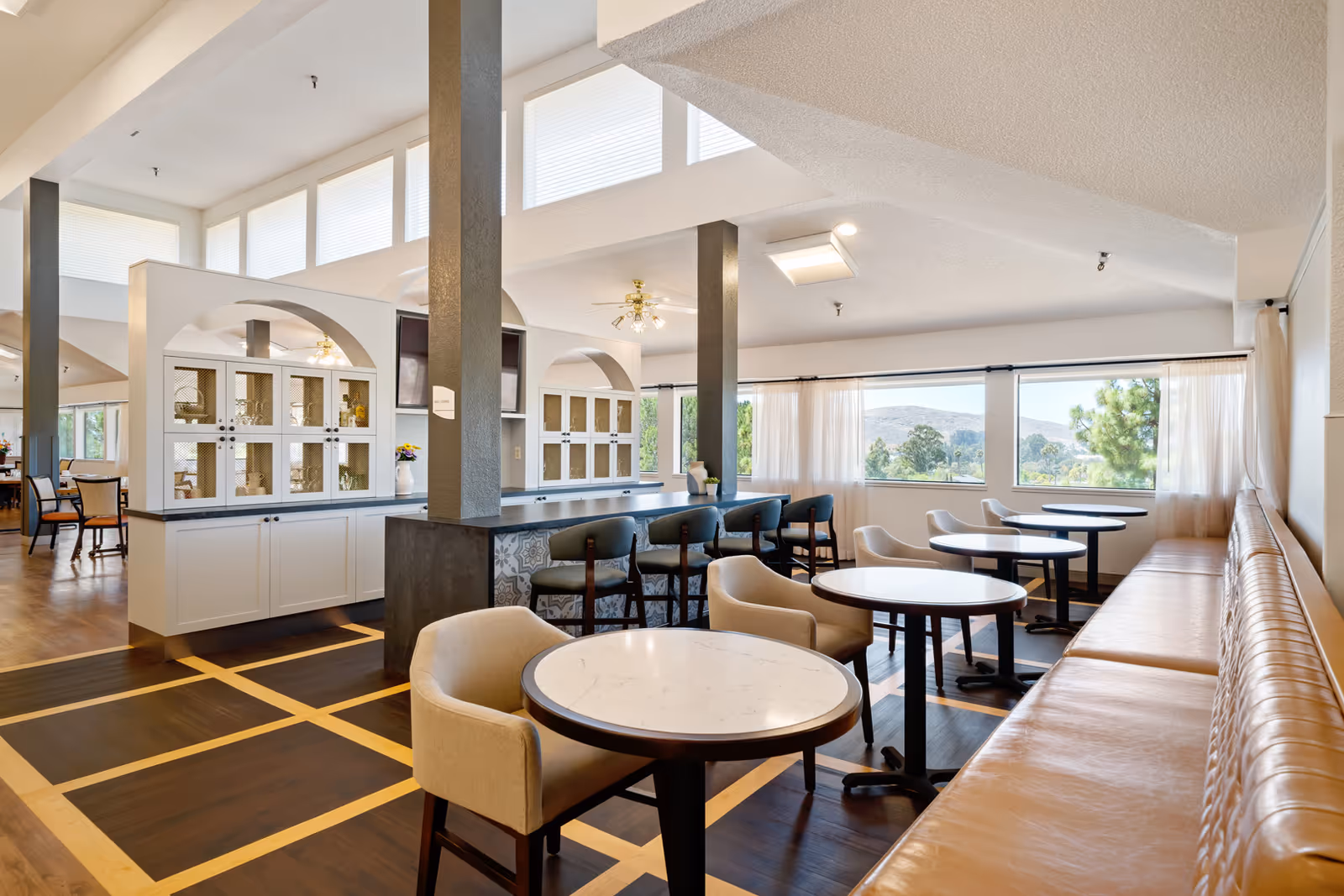 A bright and spacious dining area in a retirement community featuring round tables with beige upholstered chairs and a long tan leather bench along the windowed wall. The room has large windows with sheer curtains offering a view of trees and hills outside. There is a bar counter with high chairs and decorative cabinetry with arched openings in the background. The floor has a dark wood pattern with lighter wood inlays.