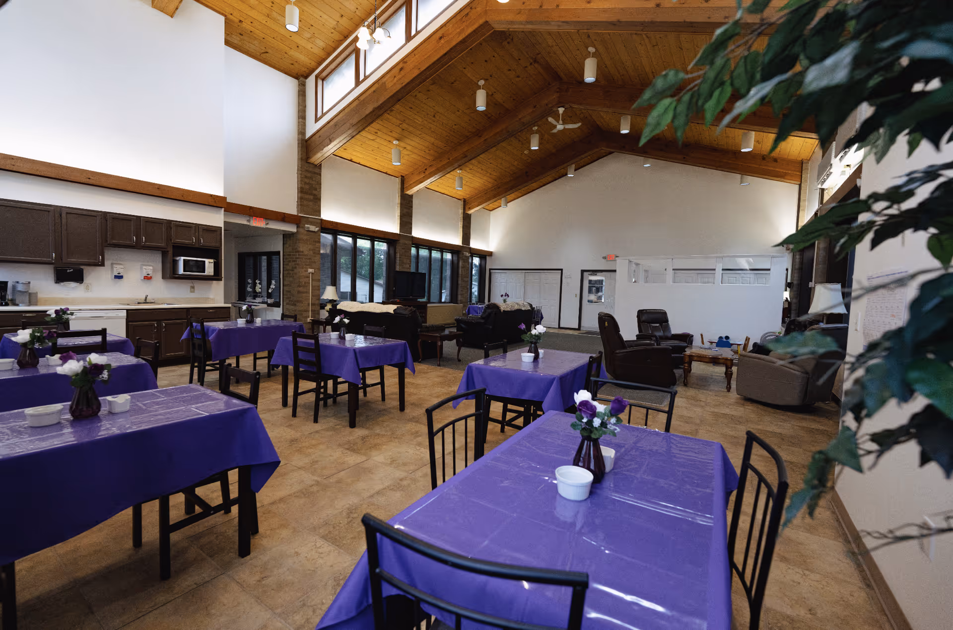 Interior view of a senior living facility dining and common area with several tables covered in purple tablecloths, each decorated with small flower vases. The room features a high wooden ceiling with exposed beams and multiple windows letting in natural light. There is a kitchenette area with cabinets, a microwave, and a sink on the left side. In the background, there are comfortable chairs and sofas arranged around a coffee table and a television.