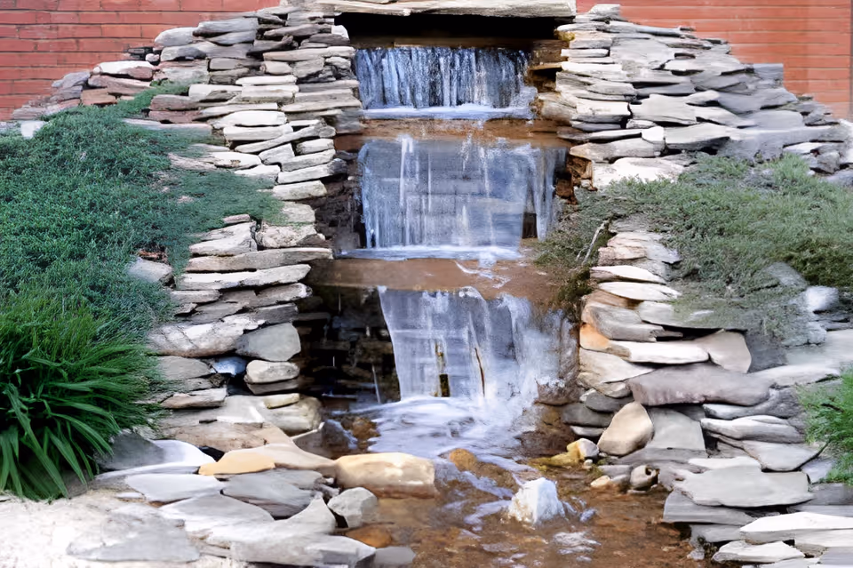 A small decorative waterfall feature made of stacked stones with water cascading down multiple levels, surrounded by green shrubs and plants, set against a red brick wall background.