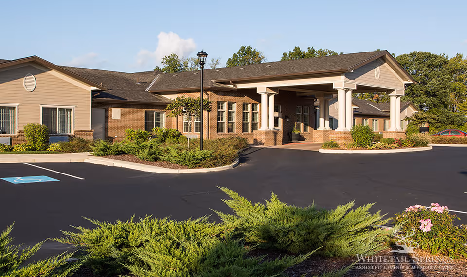 Front entrance of a single-story brick senior living facility with a covered porte-cochère, landscaped beds, and a parking area.