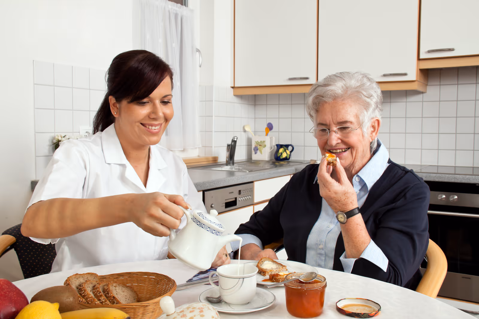 A caregiver in a white uniform is pouring tea from a teapot into a cup for an elderly woman who is smiling and holding a piece of bread with jam in a kitchen setting. The table has a basket of bread, fruit, a jar of jam, and a cup and saucer.