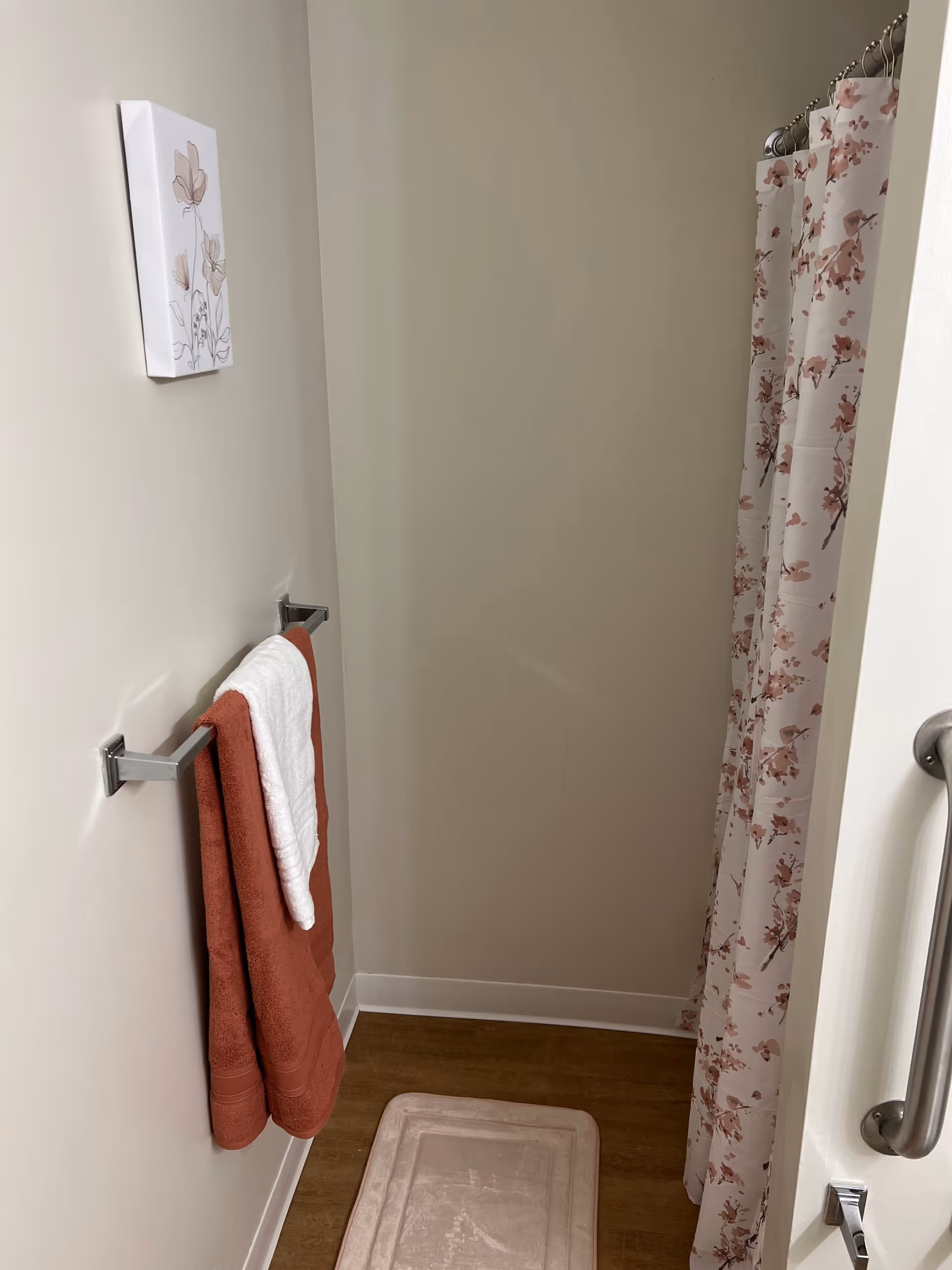 A small bathroom corner featuring a towel rack with a rust-colored towel and a white towel hanging on it. There is a floral shower curtain on the right side and a beige bath mat on the wooden floor. A simple floral artwork is hung on the wall above the towel rack.
