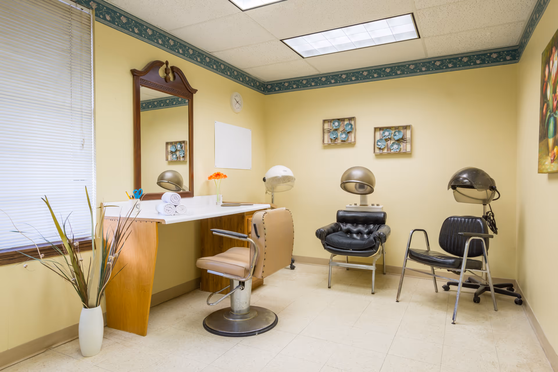 A small salon room with three vintage hair drying chairs, a large mirror above a wooden counter with rolled white towels and a vase with orange flowers, beige walls with floral border near the ceiling, a window with blinds, and decorative wall art.