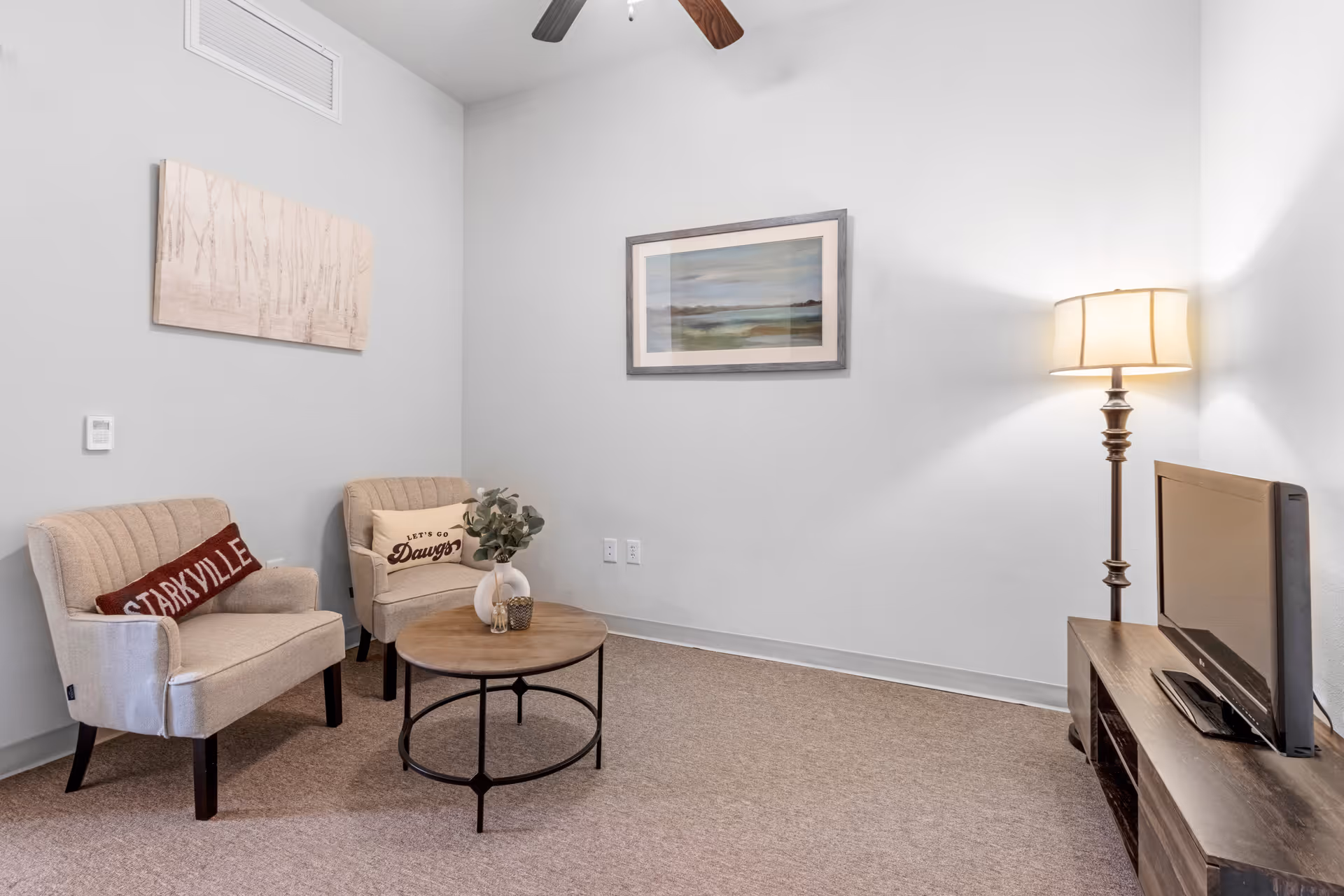 A small living room with two beige upholstered chairs, each with a decorative pillow, a round wooden coffee table with a vase and small decor, a floor lamp with a beige shade, a flat-screen TV on a wooden TV stand, and two framed artworks on white walls.