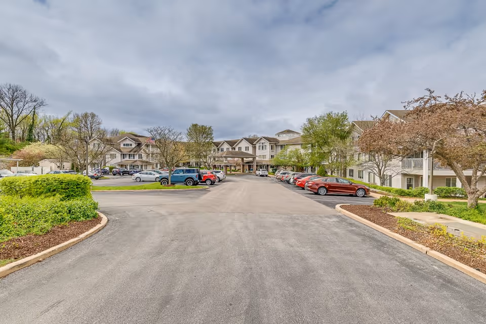Wide driveway leading to the entrance of a large senior living facility building with multiple parked cars on both sides, surrounded by trees and landscaped greenery under a cloudy sky.