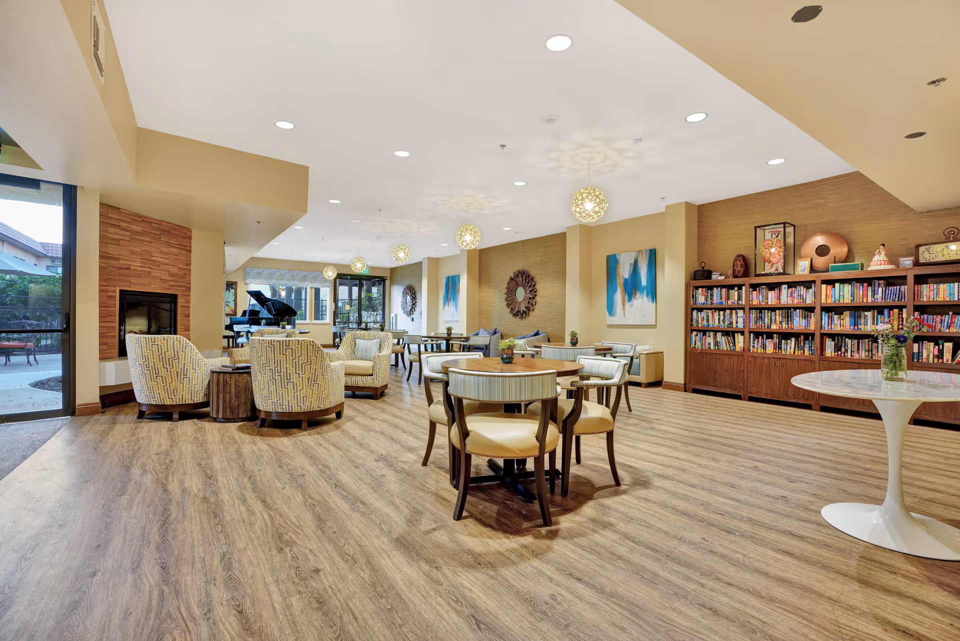 A spacious and well-lit common area in a senior living facility featuring wooden flooring, round tables with chairs, comfortable armchairs arranged near a fireplace, a bookshelf filled with books, decorative wall art, and a grand piano near large windows that open to an outdoor patio.