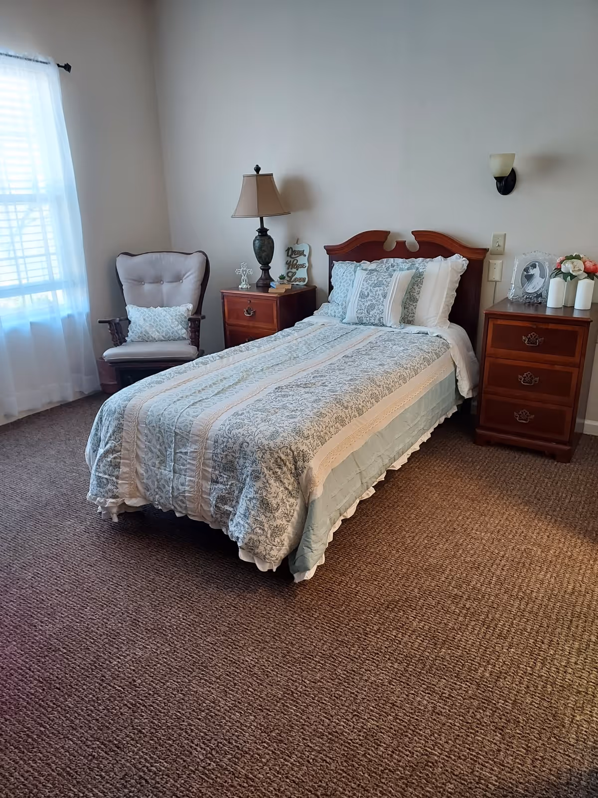 A neatly made single bed with a patterned bedspread and multiple pillows in a bedroom. The room has a cushioned armchair with a decorative pillow near a window with sheer white curtains. There are two wooden nightstands on either side of the bed, one with a table lamp and decorative items, and the other with a framed photo and a vase with flowers. The walls are plain and light-colored, and the floor is carpeted.