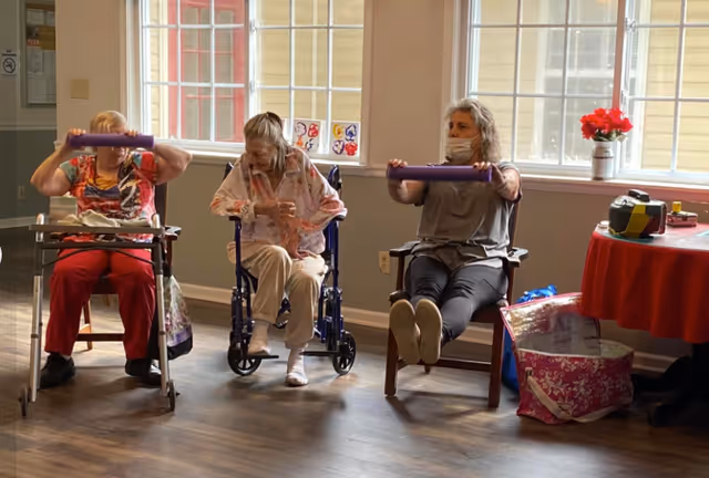 Three elderly women participating in a seated exercise session in a room with large windows. Two women are holding purple exercise bars, while the woman in the middle is seated in a wheelchair. The room has wooden flooring, a red table with a flower vase, and some personal items nearby.