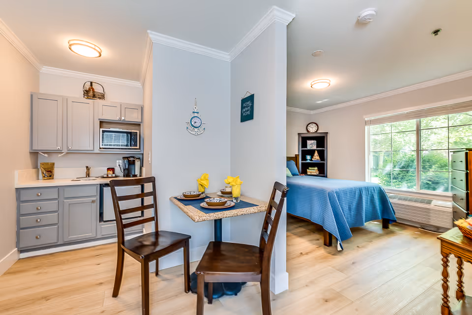 A cozy senior living studio apartment featuring a small kitchen with gray cabinets, a microwave, and a coffee maker. Adjacent to the kitchen is a small dining table set for two with yellow napkins. The sleeping area has a bed with a blue bedspread, a bookshelf, and a large window letting in natural light. The room has light-colored wooden flooring and soft lighting fixtures on the ceiling.
